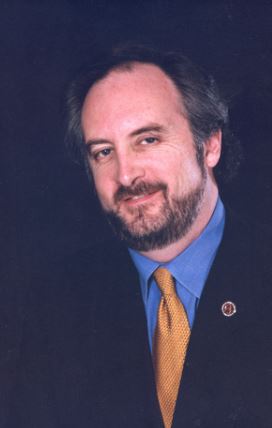 A man with a short beard and receding hairline looks toward the camera with a gentle smile. He is wearing a navy suit jacket, a blue shirt, and a golden-yellow tie with a small round pin on his lapel. The background is solid black, giving the image a formal tone.