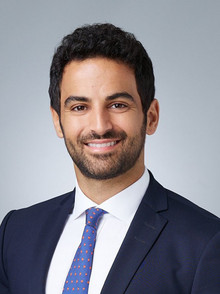 A young man with dark curly hair and light stubble smiles confidently. He wears a white shirt, a navy suit, and a blue tie with red dots. The backdrop is a smooth gradient from light to medium gray, enhancing the professional tone of the photo.