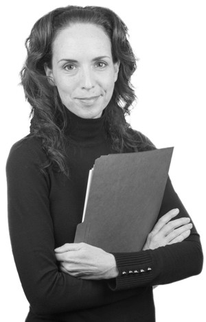 Black-and-white portrait of a woman with long, wavy hair, wearing a black turtleneck. She is smiling slightly and holds a folder across her chest with both arms crossed.